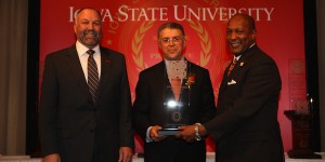 Esmail Zirakparvar holds a large, glass award on stage next to Jeff Johnson and Steven Leath with an Iowa State backdrop