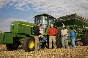 An agronomist, beef specialist, and FSA employee meet with two farmers in a cut corn field by a John Deer tractor and auger cart