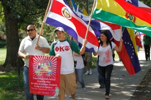 Multicultural Students walk on a sidewalk holding a sign and proudly carrying their country's flags.