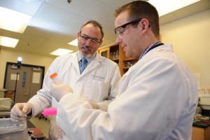 Alum Lucas Carlstrom helps Matthew Ellinwood in his lab. They both wear white lab coats and safety glasses while examining substances in a tube.