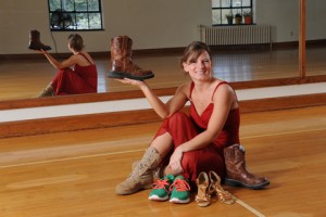Jenny English sits in an ISU dance studio surrounded by multiple pairs of shoes. She holds up a cowboy boot.