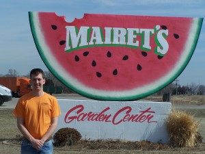 Shane Mairet stands in front of a sign for Mairet's Garden Center that is shaped like a watermelon slice
