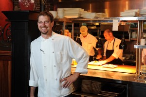 Kevin Rettig leans against a post smiling in his chef coat with food preparation in the background.