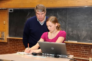A professor stands behind a student helping her prepare for a presentation