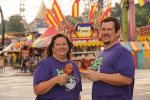 Two people eat turkey legs at the Iowa State Fair. A carnival is in the background.