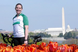 Sarah Low wears a USDA shirt standing next to a bike by a tulip garden with the city of Washington DC in the background.