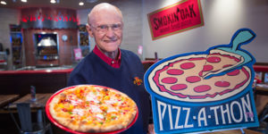 Retired affiliate instructor, Eldon Weber, holds out a pizza by a Pizz-A-Thon sign