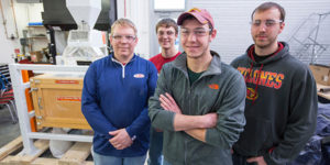 A group of four students pose wearing safety glasses with a grain sifter in the background