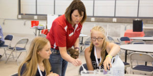 A 4-H leader leans over two girls sitting at a table working on a project
