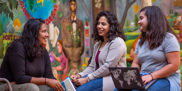 Carver Award winners Valeria Cano Camacho, Estefany Argueta and Megan Kemp visit between classes in Horticulture Hall seating in front of a colorful mural by Sticks featuring George Washington Carver.