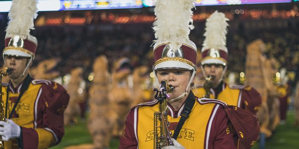 Dawn Henderson plays saxophone in Cyclone marching band uniform on Jack Trice field at night with stadium lights glowing behind her.