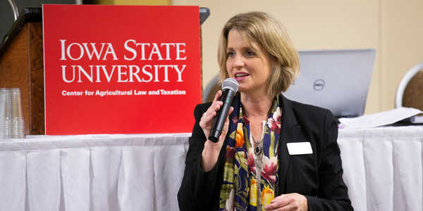 Kristine Tigren speaks into a microphone while standing in front of an Iowa State University sign while presenting for the Center for Ag Law and Taxation