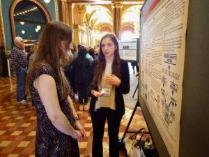 undergrad talks with a visitor in front of a poster about her research in the rotunda of the Iowa State Capitol building