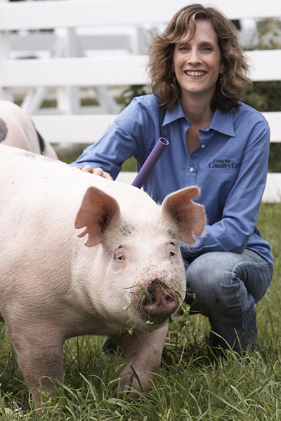 Betsy Freese wearing a blue button up shirt and kneeling next to a pink pig