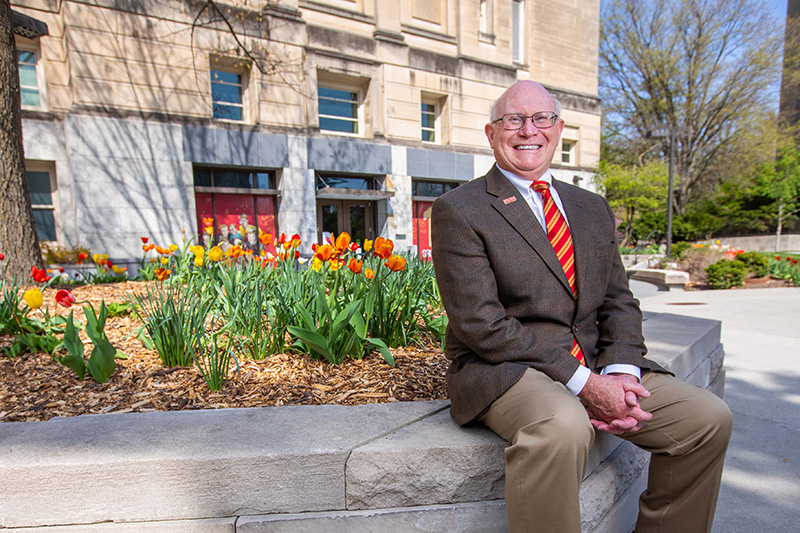 Joe Colletti, pictured sitting on a stone wall with blooming red and yellow tulips behind him.