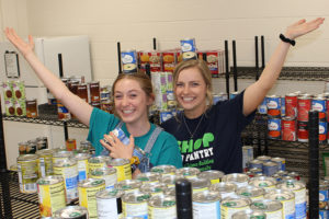 Caroline Stokes and Belinda Heckman standing with their arms up in the air, surrounded by shelves filled with canned food.